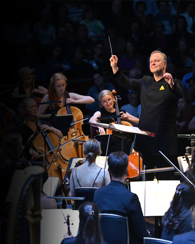 A man conducting a group of musicians, wearing formal black concert attire, playing on string instruments, with an audience in the background.